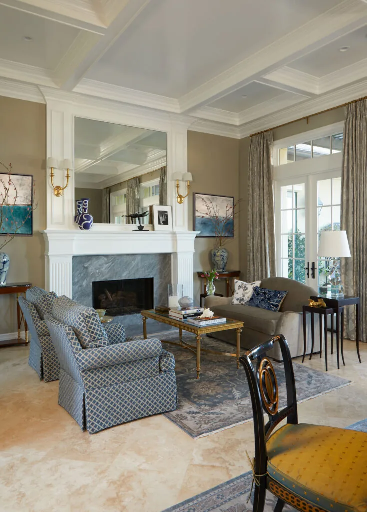 A traditional living room with a coffered ceiling, a marble fireplace and soft grey armchairs