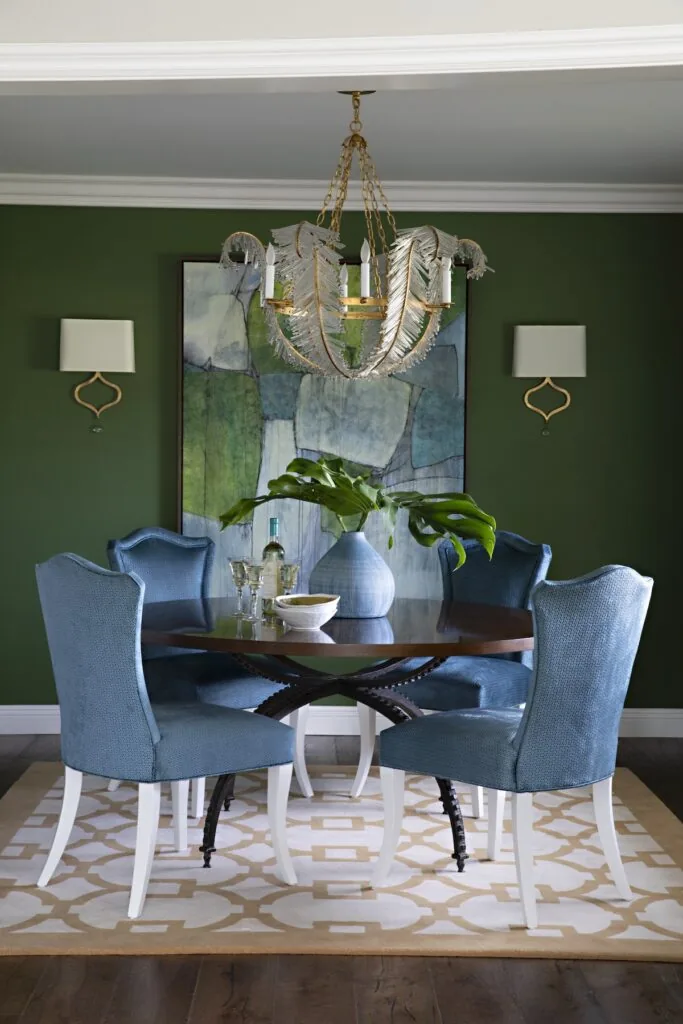 A dining room with deep green walls, blue upholstered chairs and a glass bead chandelier