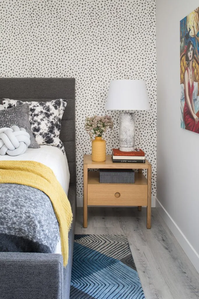 A bedroom with a grey upholstered bed, a yellow throw and a blue patterned rug